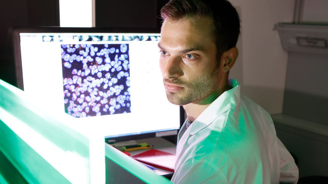 A scientist sits in front of a high-throughput microscope. The screen behind him shows cells.