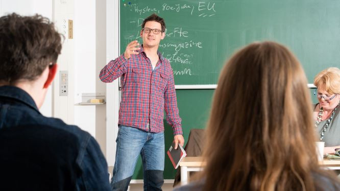 Studierender steht vor anderen Studierenden vor der tafel und trägt etwas vor